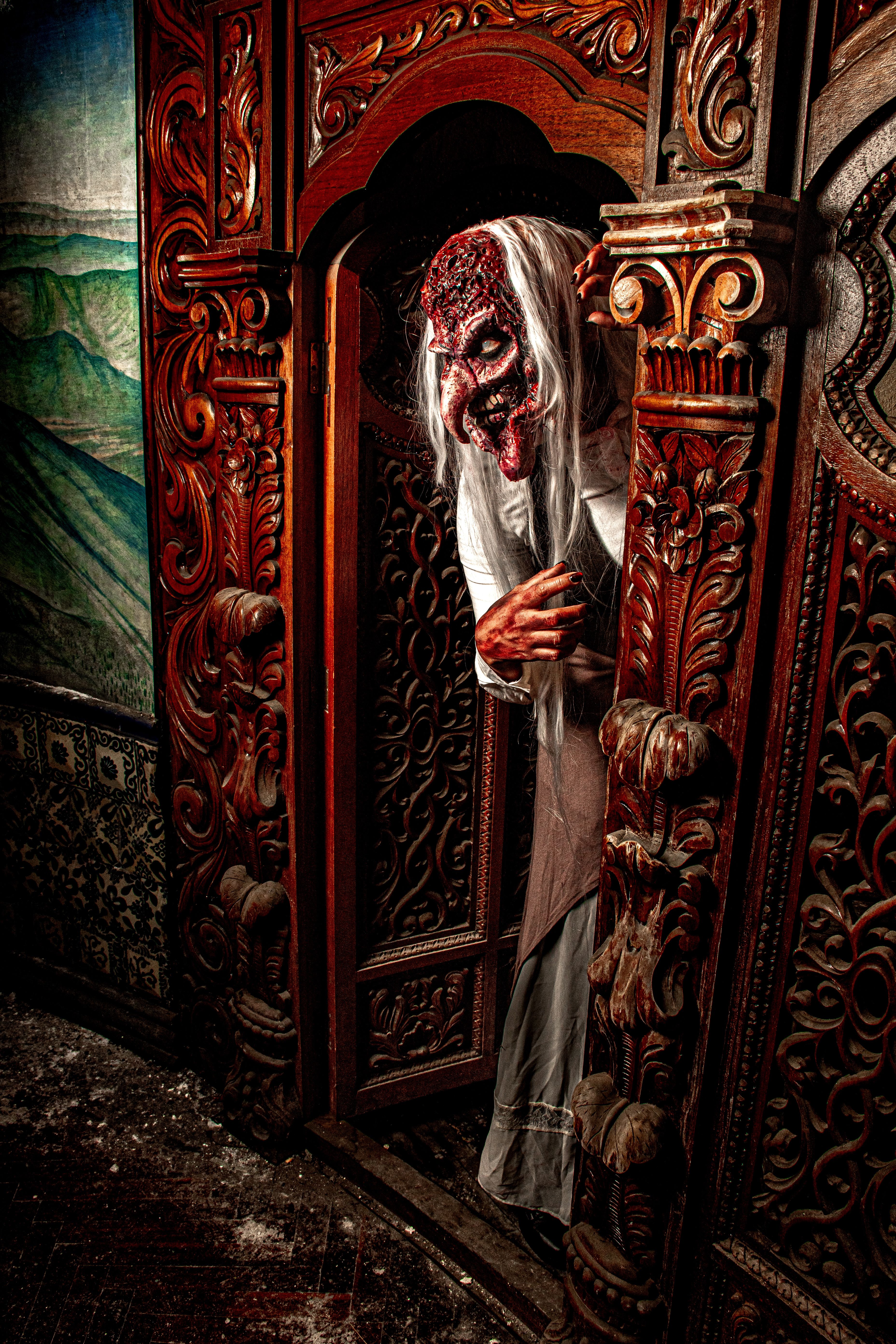 Terrifying figure with mask leans out from ornate wooden doorway, creating a haunting atmosphere.