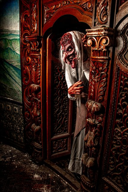 Terrifying figure with mask leans out from ornate wooden doorway, creating a haunting atmosphere.