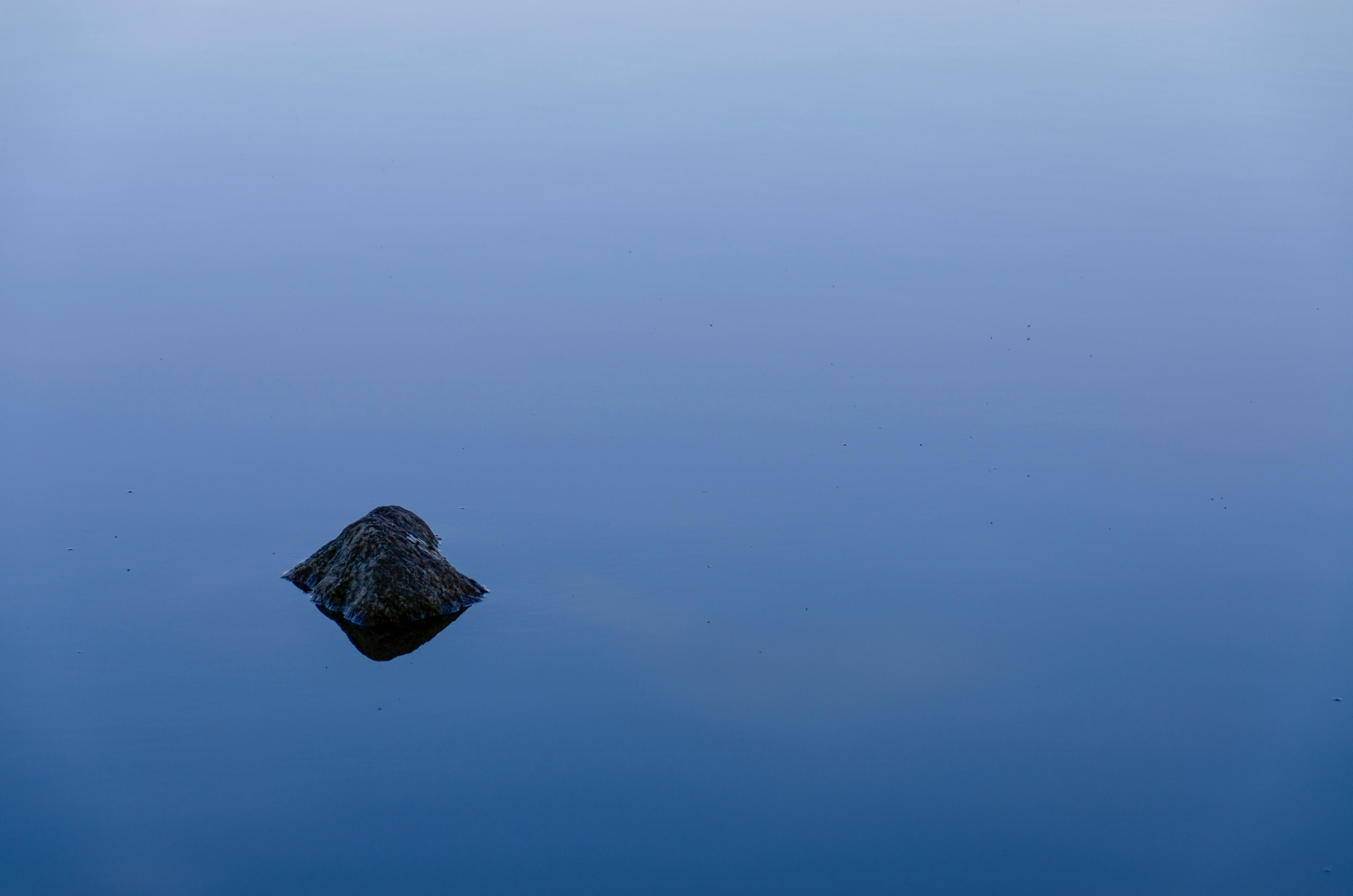 Free A lone rock reflects on a clear, tranquil water surface creating a minimalistic and peaceful scene. Stock Photo
