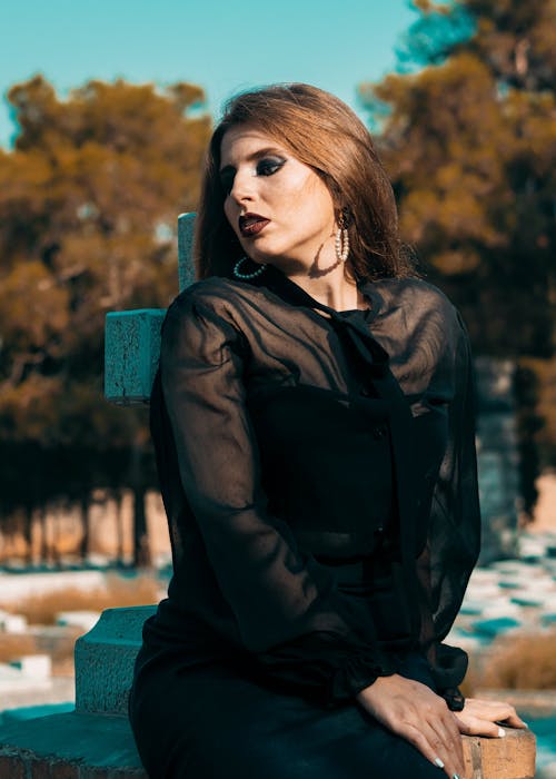 Free Thoughtful woman dressed in black clothes sitting on concrete headstone while grieving about death in cemetery Stock Photo