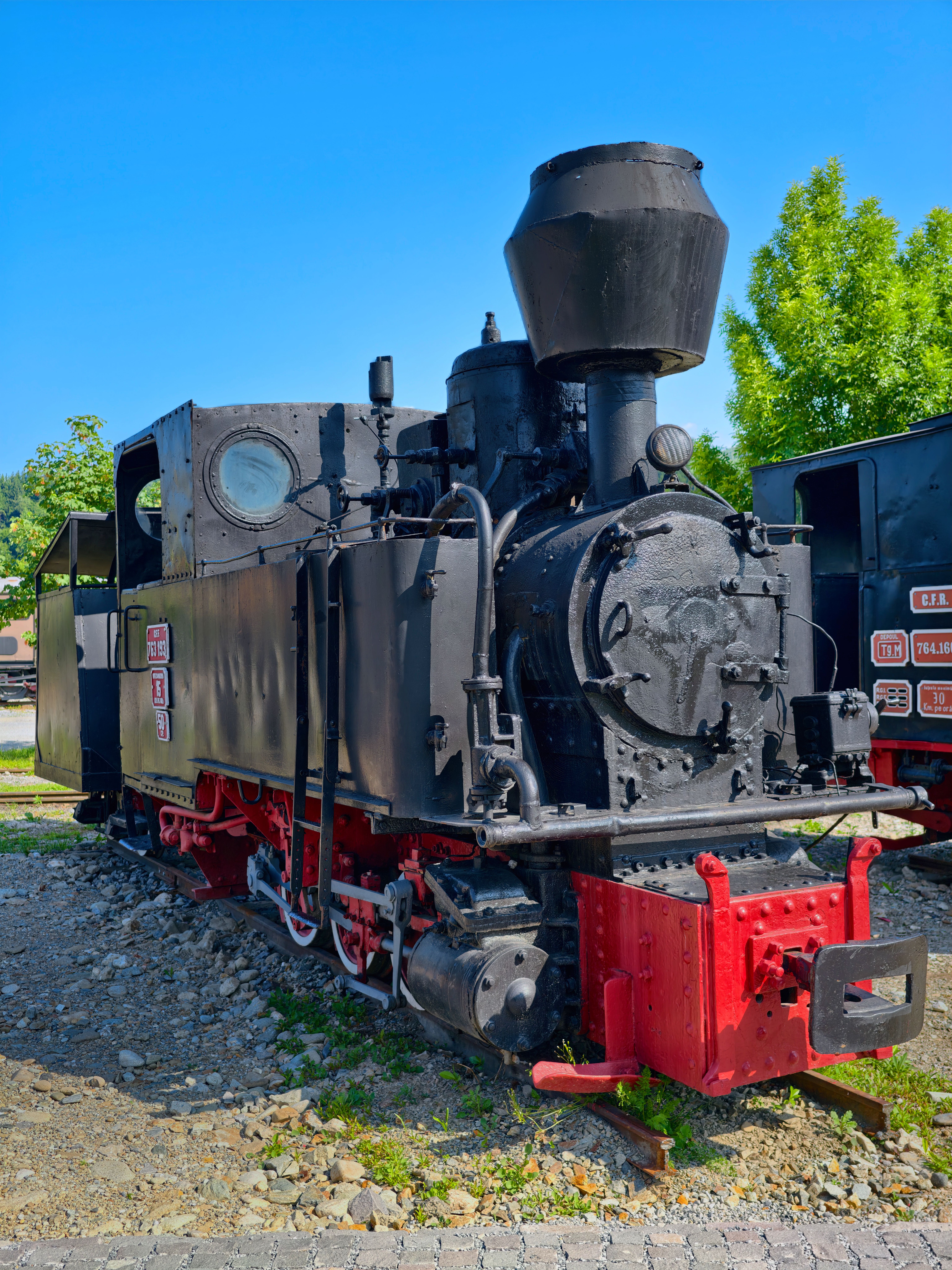 Fotomural Clásica locomotora de vapor negra de principios del siglo XX expuesta al aire libre bajo un cielo azul brillante.