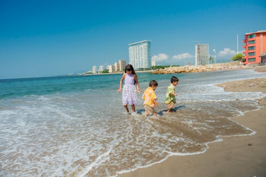 Three children play joyfully on a sunny beach with a city skyline in the background.