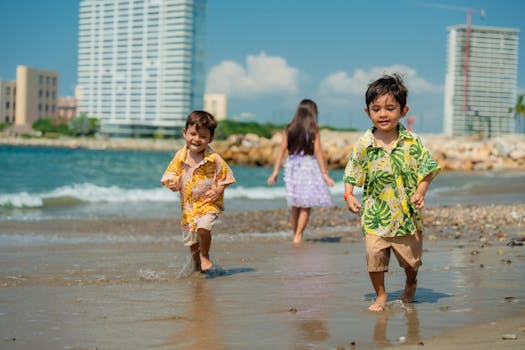 Three children playing happily on a sunny beach with the sea and buildings in the background.