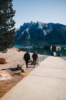 Two hikers walking near a scenic Canadian lake with mountains and clear sky in the background.