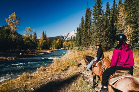 Two people enjoy horseback riding along a scenic Canadian river with forest and mountains.