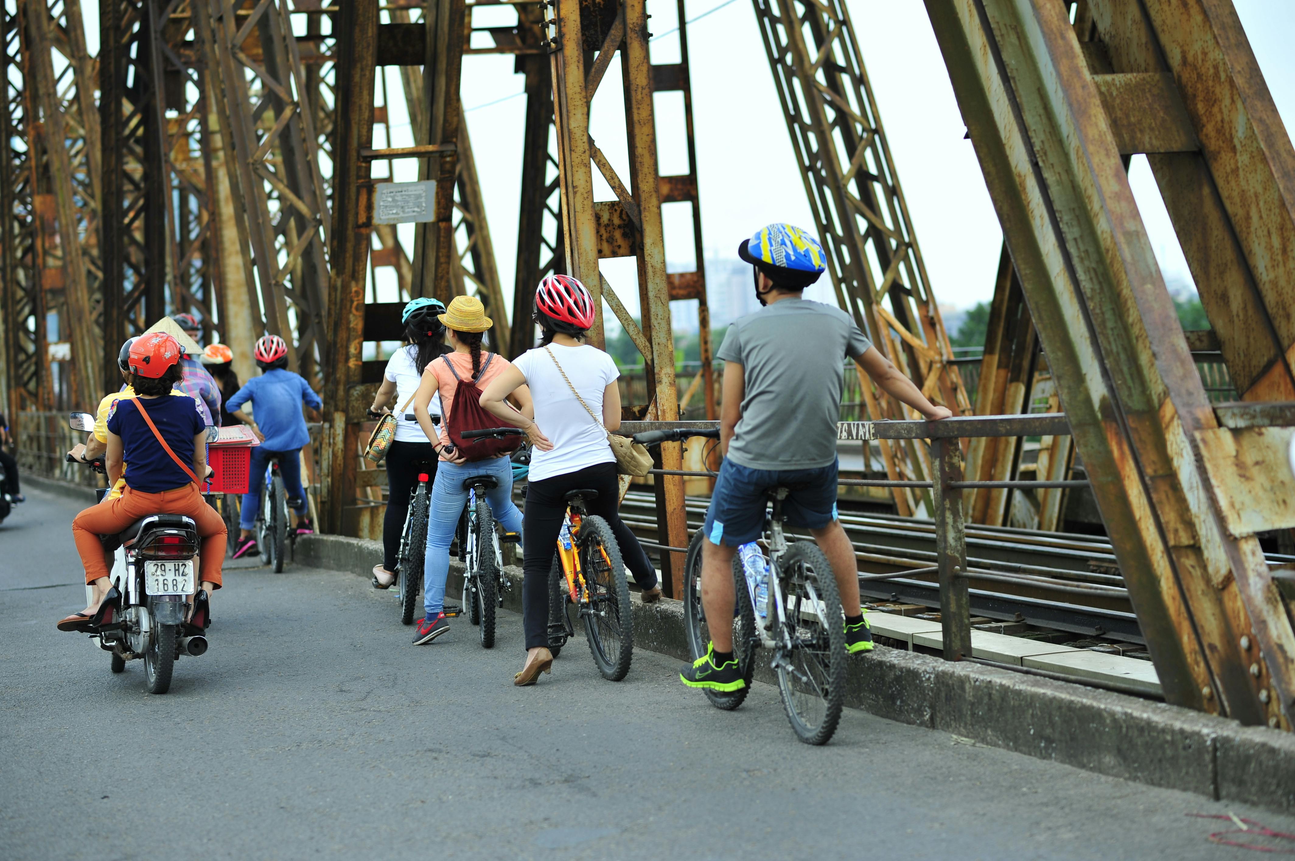 People on bicycles and a motorbike crossing a rusty bridge with iron structures.