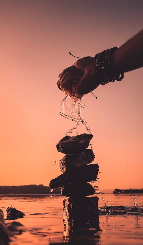 Silhouette of unrecognizable person splashing water from pond on stones stacked on top of each other during sunset