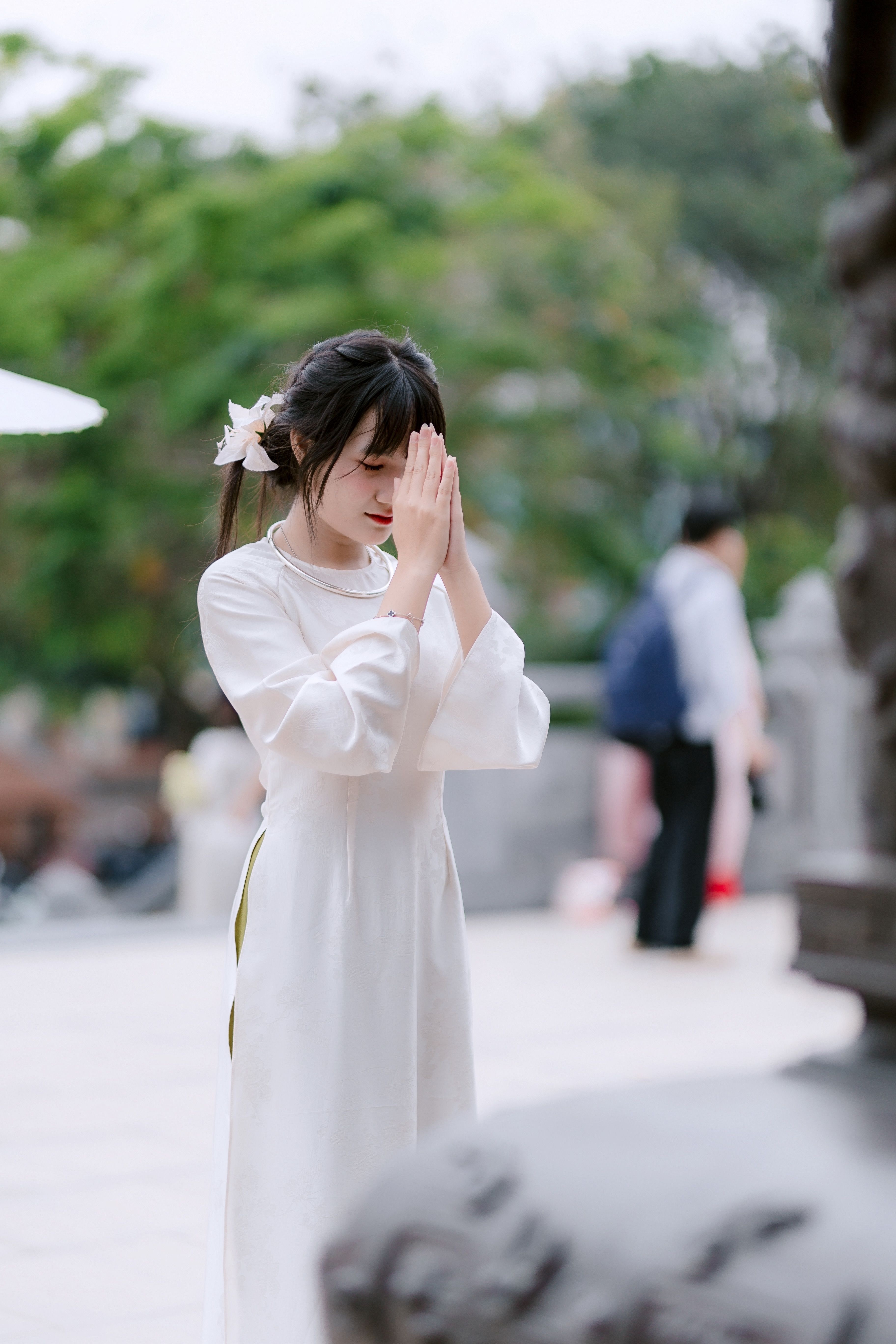 A woman in a white traditional dress offering a prayer outdoors in a peaceful setting.