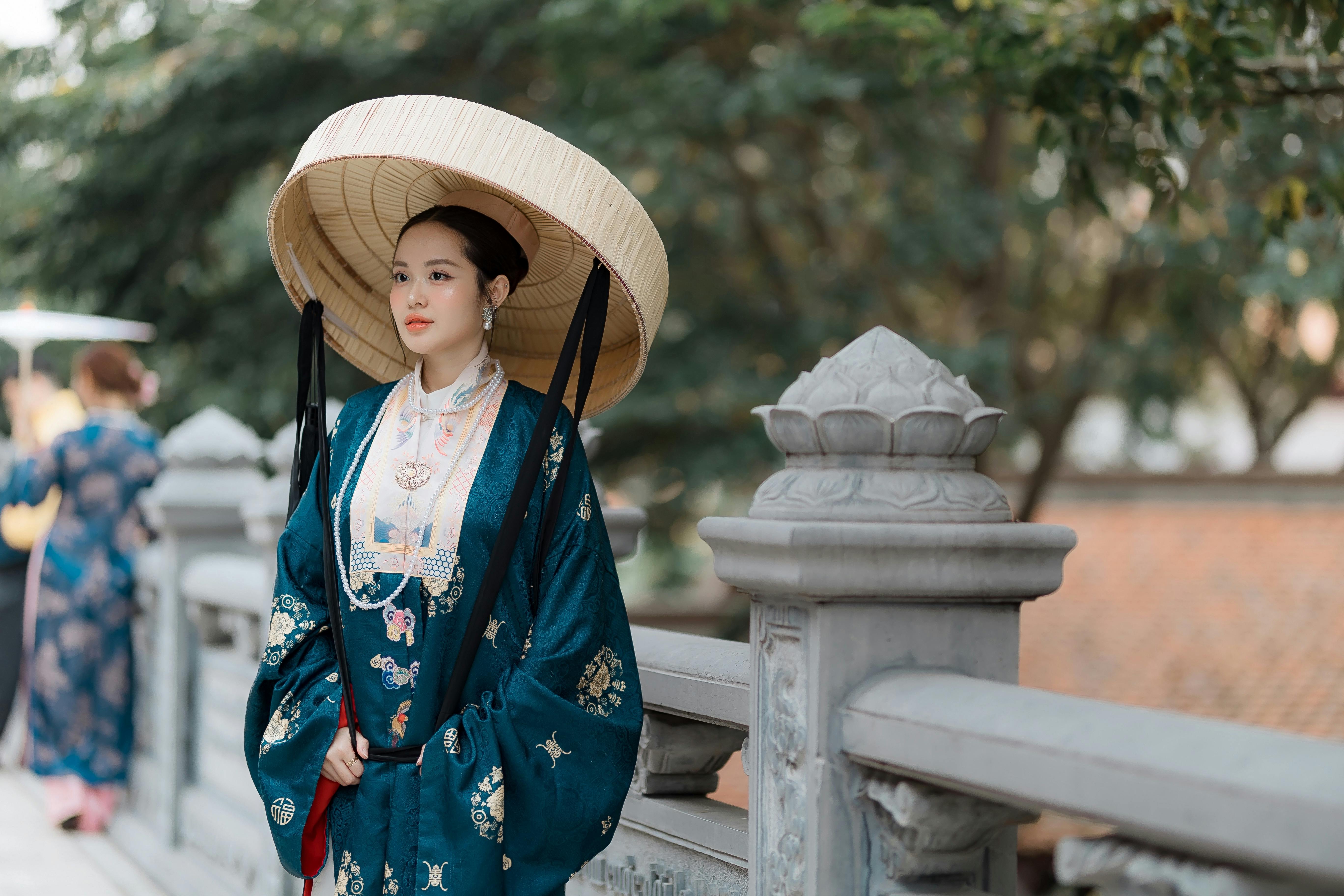 bisht robe - Elegant woman in traditional attire with a straw hat, outdoors on a historical bridge.