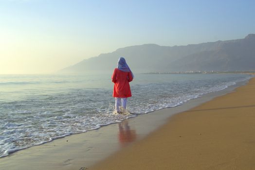 A woman in red enjoys a peaceful morning at As Sifah Beach, Oman, with misty mountains as backdrop.