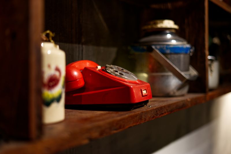 A vintage red rotary telephone sits on a rustic wooden shelf indoors, surrounded by antique items.