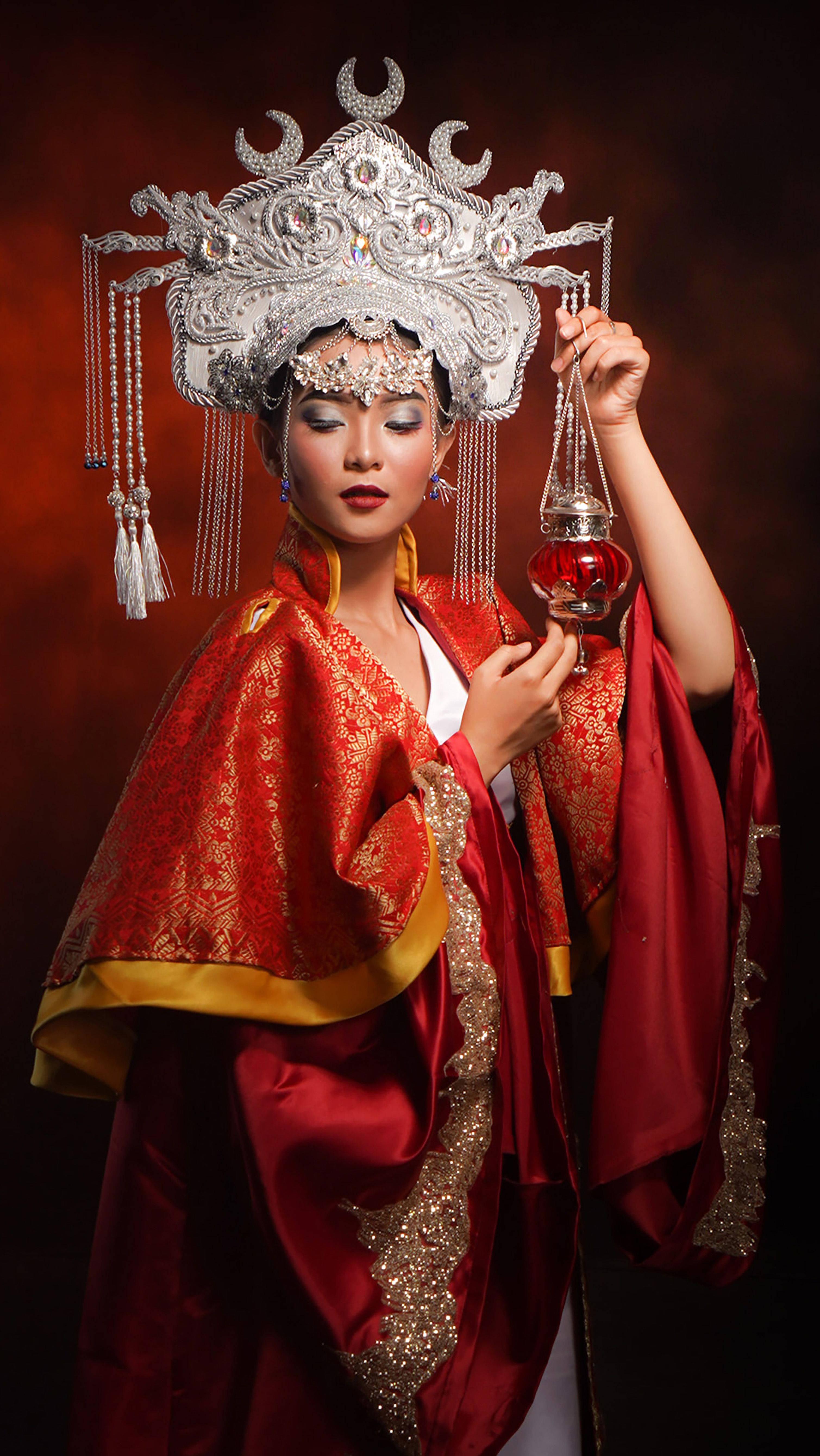 Elegant woman in traditional Asian attire and intricate silver headpiece holding a teapot.
