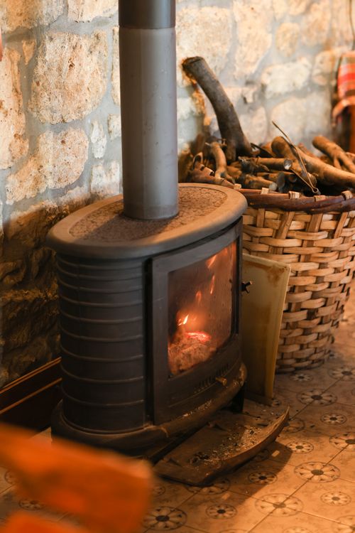 Free Warm and rustic indoor scene featuring a lit wood stove by a stone wall with a basket of firewood. Stock Photo