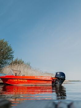 A vivid red motorboat reflecting in a serene lake, surrounded by nature under a clear sky. — photo by Andis Bāliņš