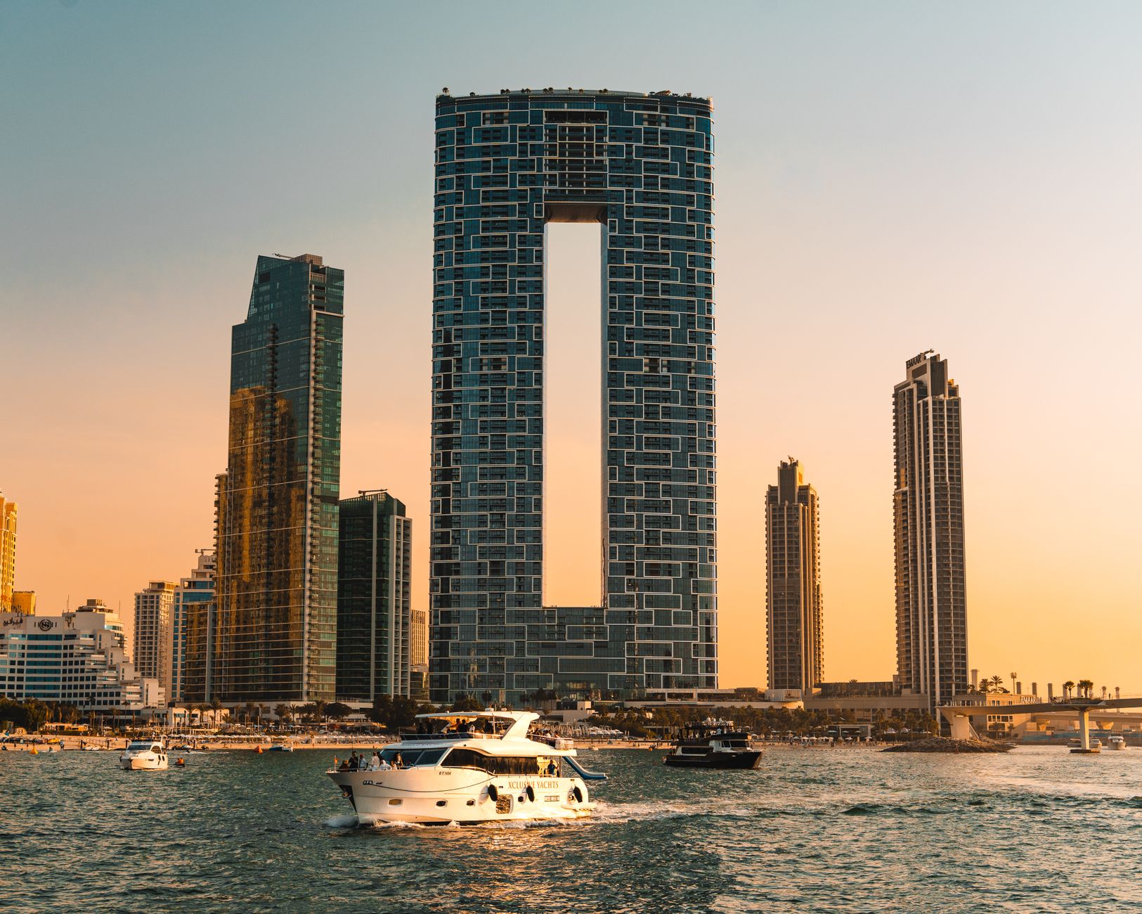 A luxury yacht cruises against the modern architectural skyline of Dubai at sunset.