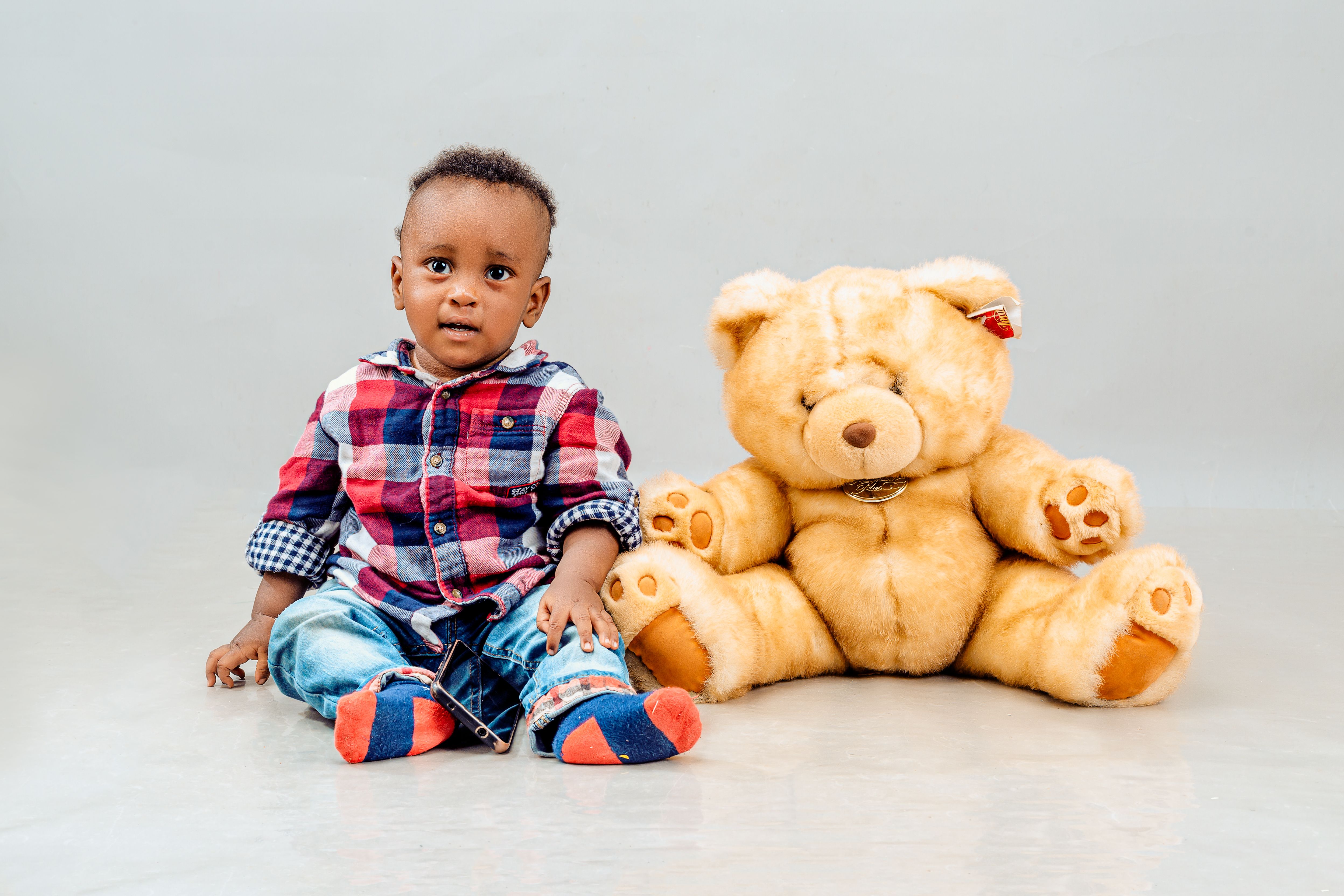 Cute baby in plaid shirt sitting with a fluffy teddy bear in a bright studio.