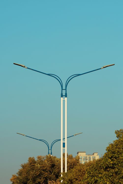 Free Symmetrical street lamps extend into a clear sky with trees and a building in the background. Stock Photo