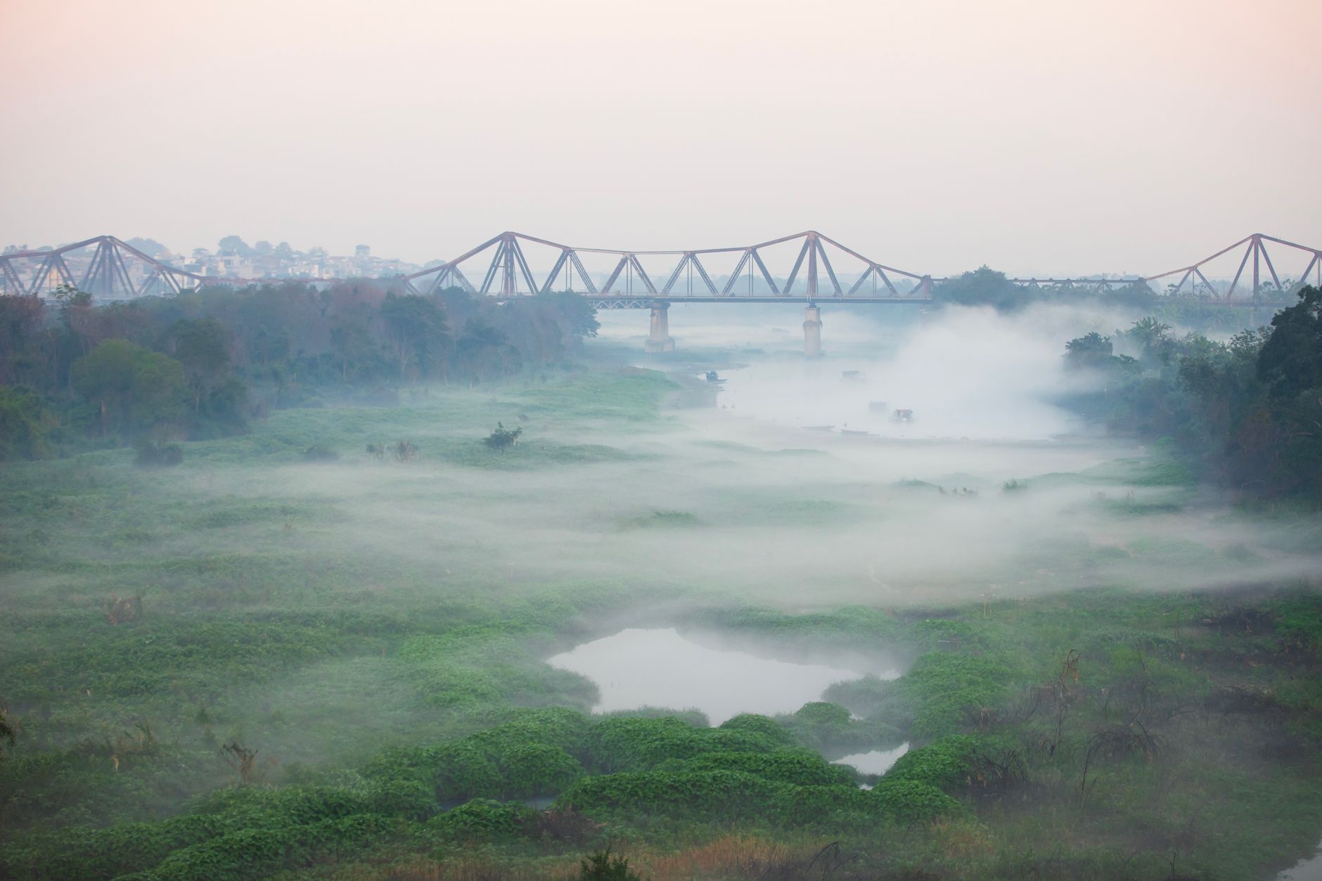 A serene misty morning view of Long Bien Bridge over lush greenery in Hanoi, Vietnam.