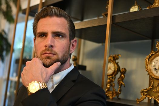 Portrait of a thoughtful man in formal wear with vintage clocks and gold watch indoors.