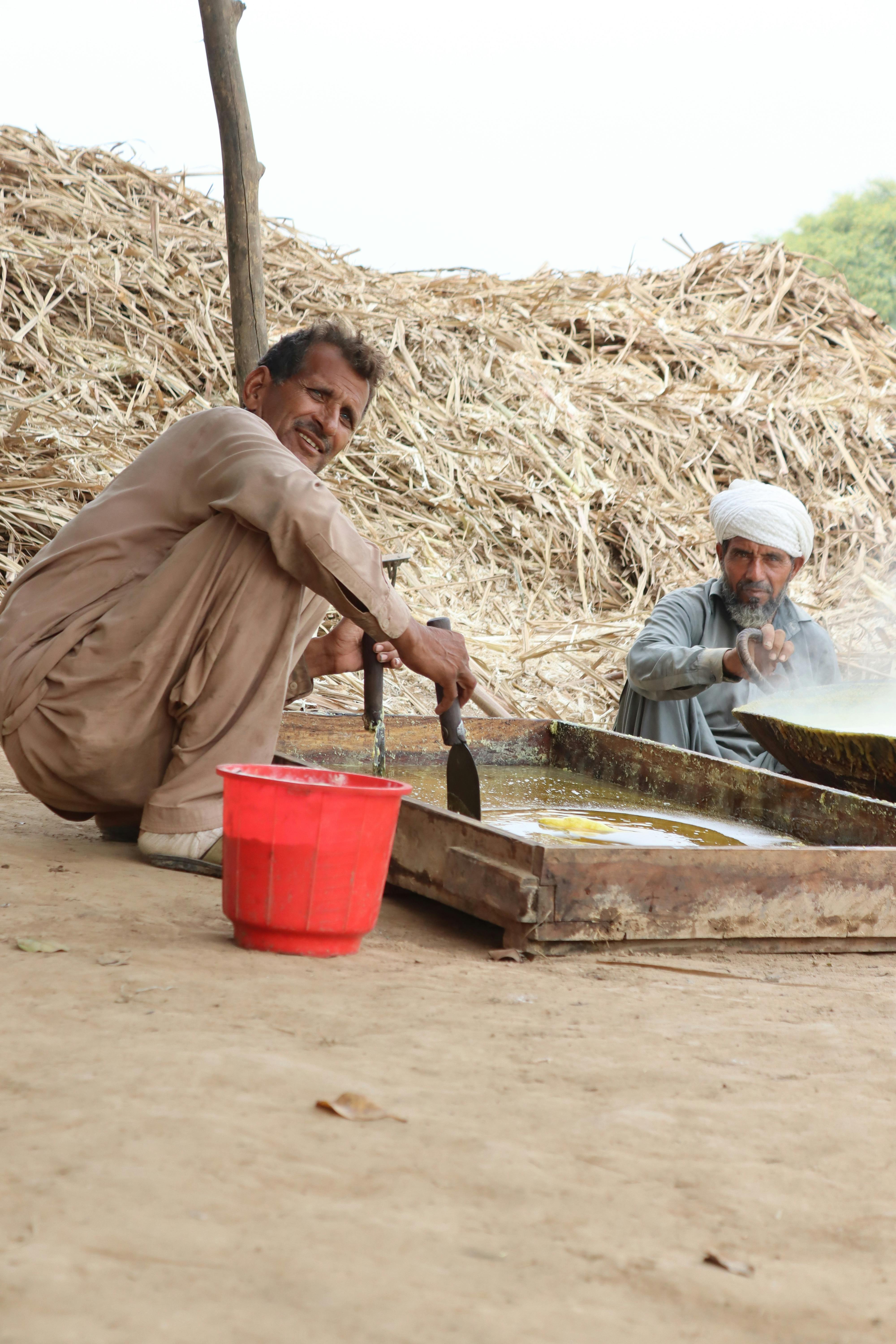 Two men cooking outdoors in a rural setting surrounded by hay and using traditional methods.