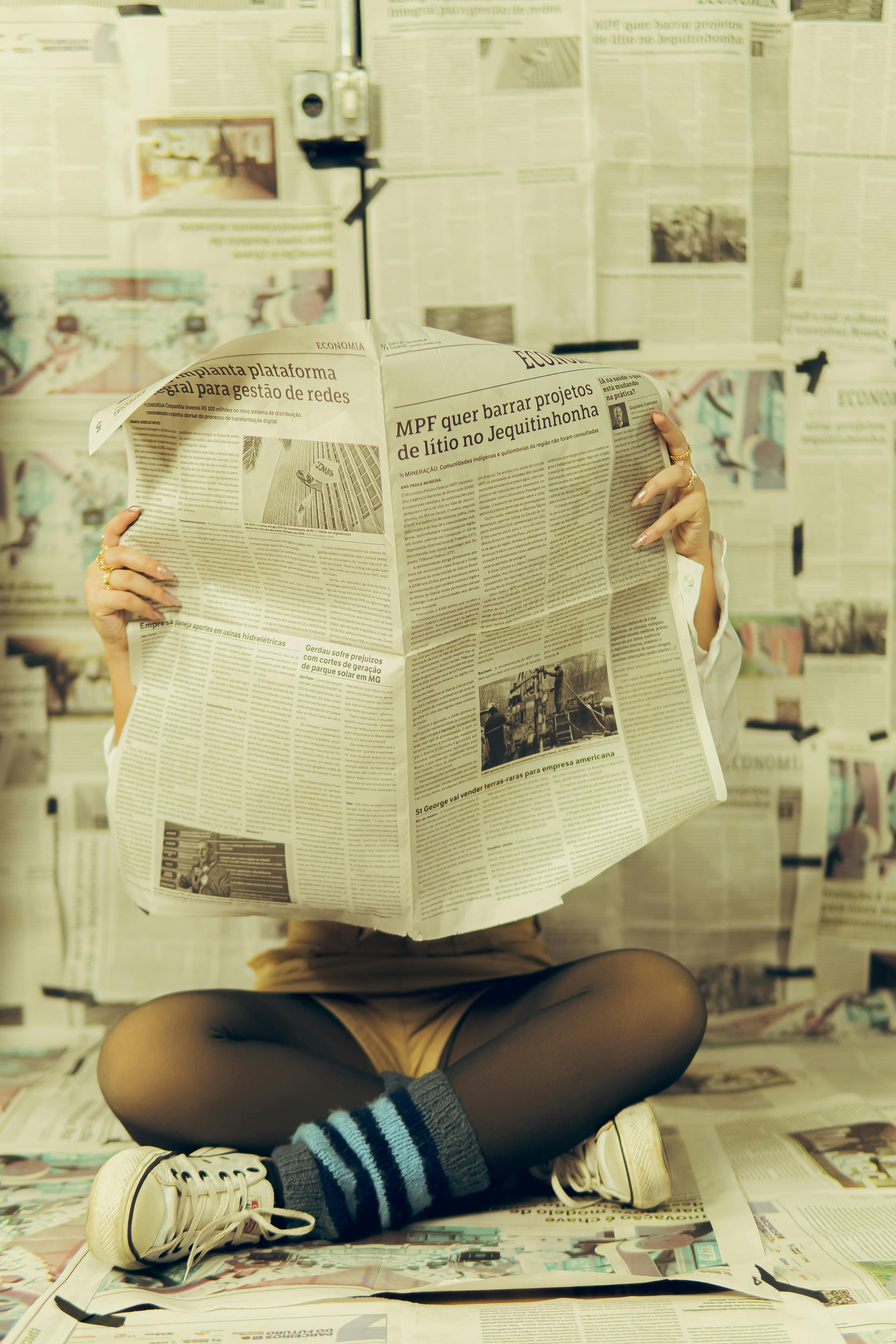 Individual sits on floor reading a newspaper, surrounded by newspapers as wallpaper.
