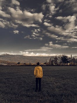 一个穿着黄色夹克的人独自站在草地上，凝望着远处山脉上那戏剧性的云层