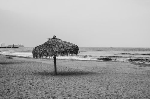 Free A serene black and white photograph of a beach with a straw umbrella and gentle waves. Stock Photo