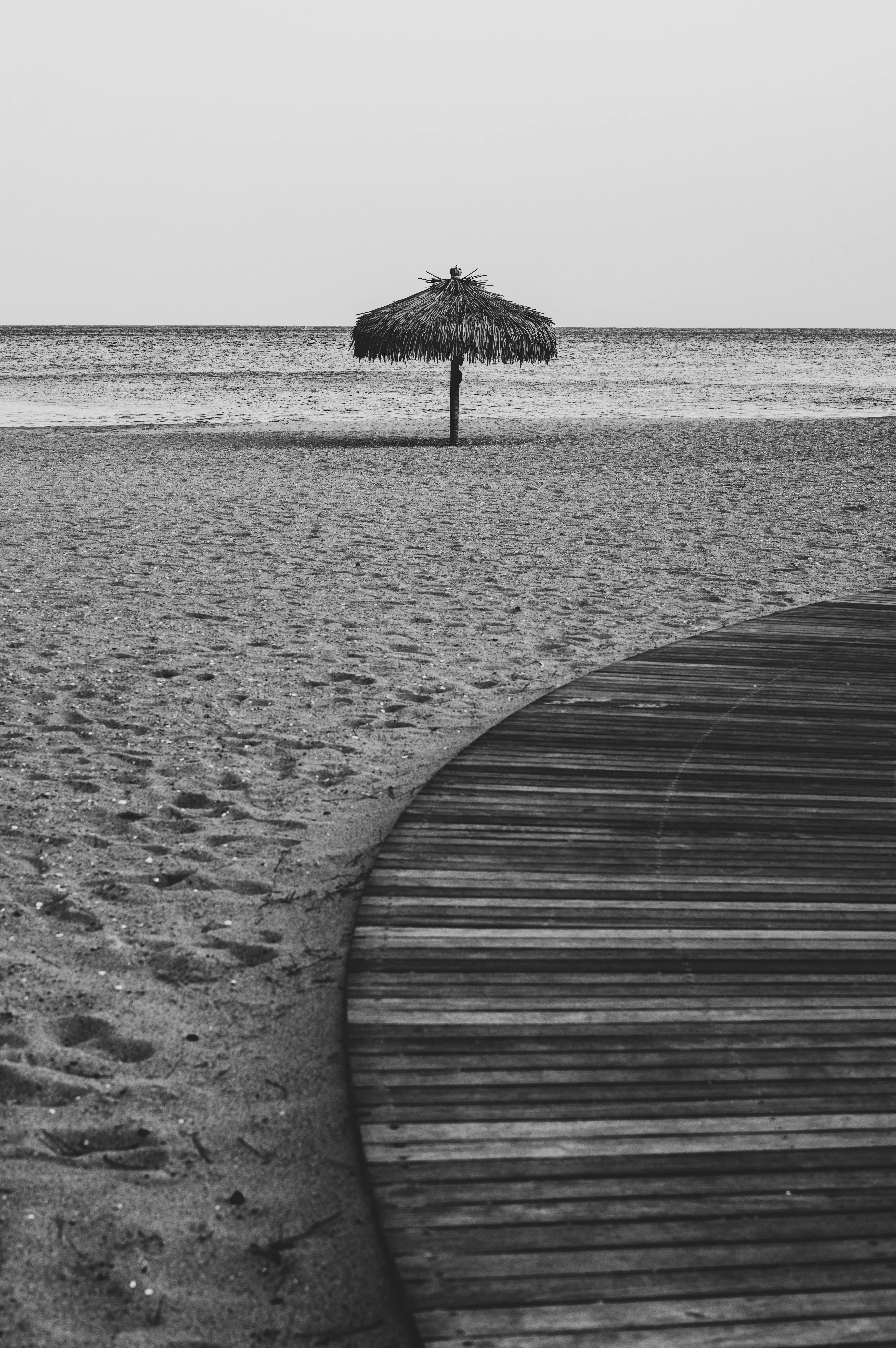 Black and white photo of an empty beach with a straw umbrella and wooden walkway.