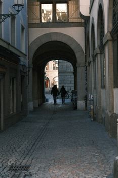 Silhouette of two people walking through a European-style archway in a narrow cobblestone alleyway.