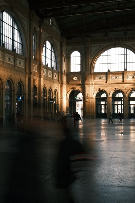 Large hall of a historic train station with arched windows and people moving in dim light.