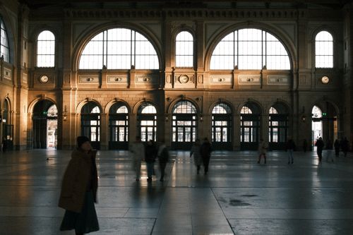 Free Interior view of a classic train station with large arched windows and blurred figures passing through. Stock Photo