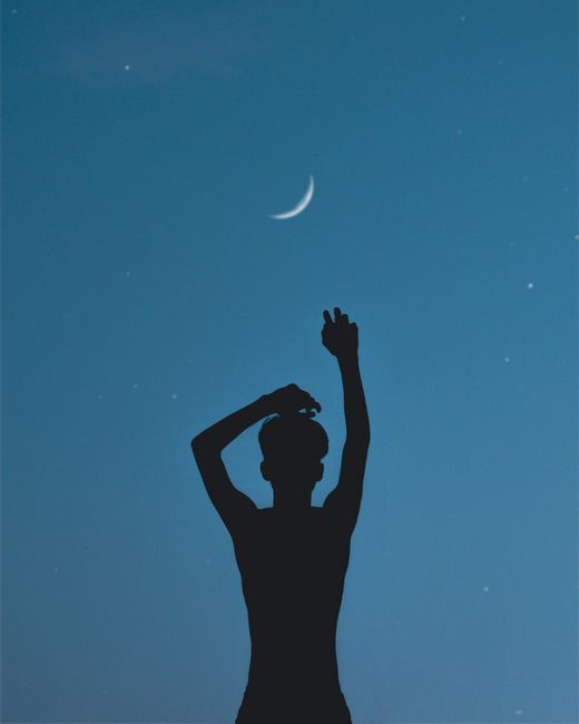 Silhouette of a person raising hands under the crescent moon in a clear night sky.
