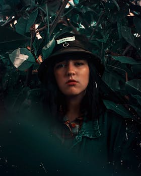 一位年轻女子头戴帽子，在郁郁葱葱的深绿叶丛中摆出姿势，散发出神秘的氛围