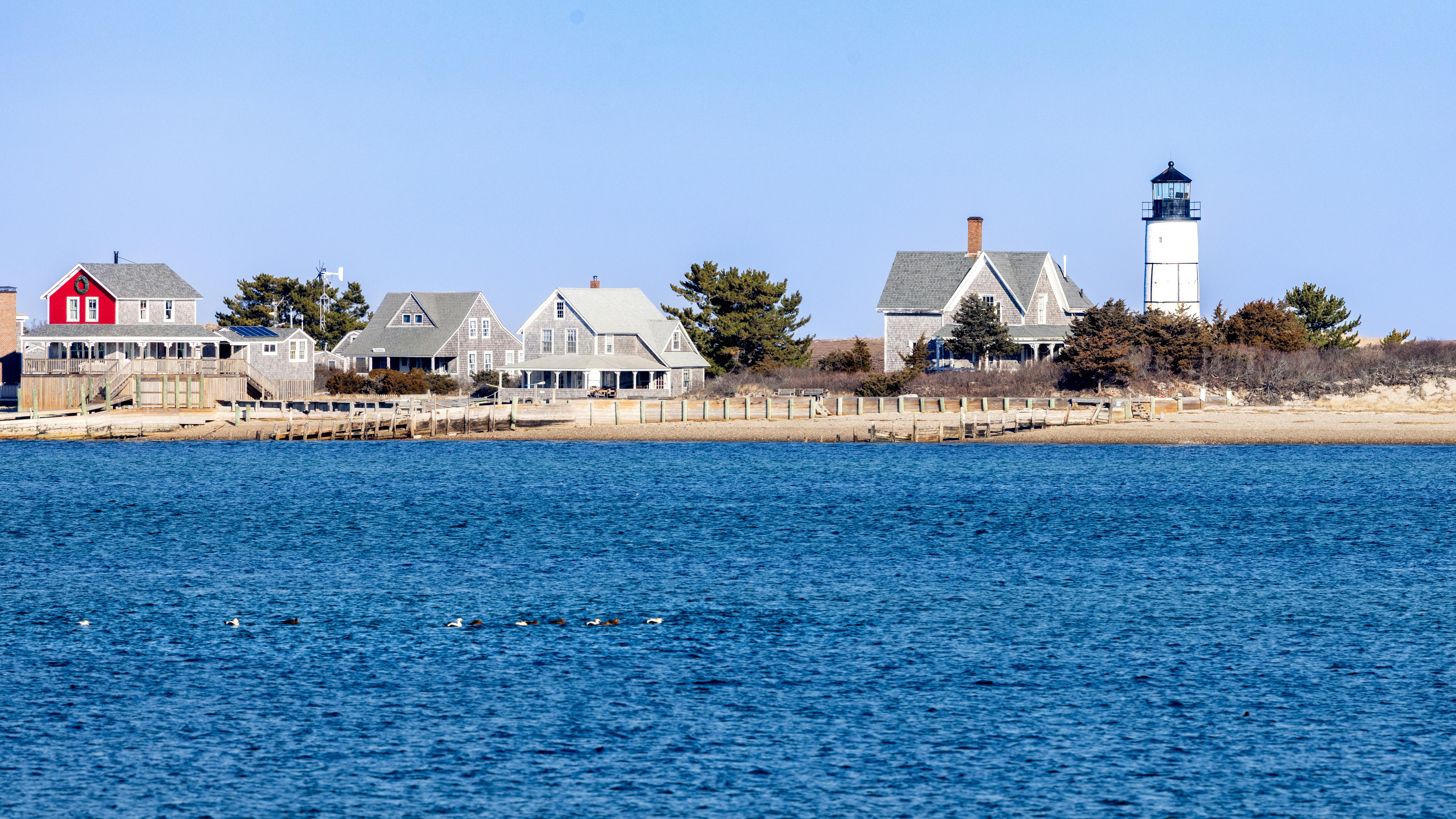 Picturesque Cape Cod lighthouse with traditional homes by the shoreline under a clear sky.