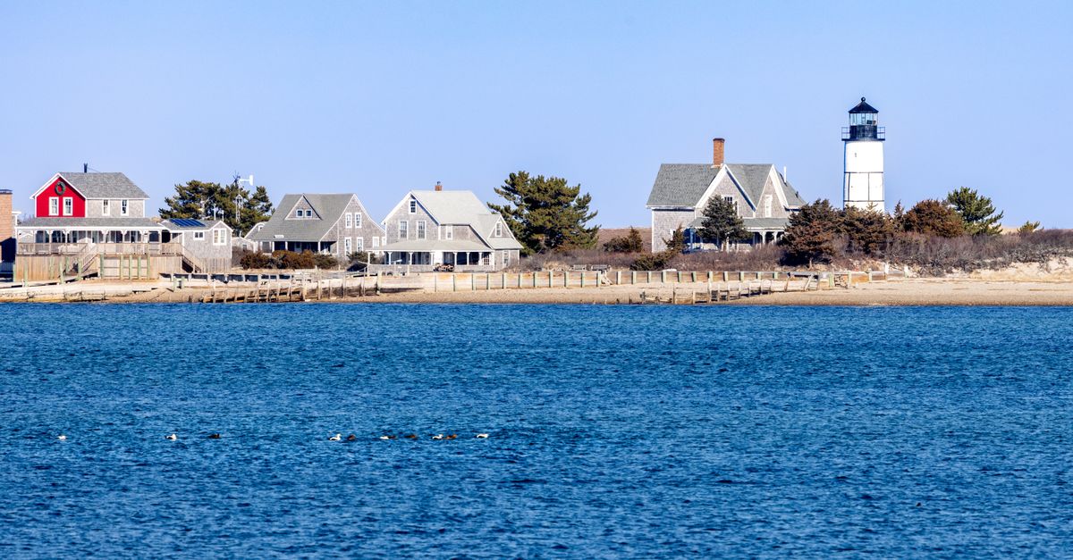 Picturesque Cape Cod lighthouse with traditional homes by the shoreline under a clear sky.