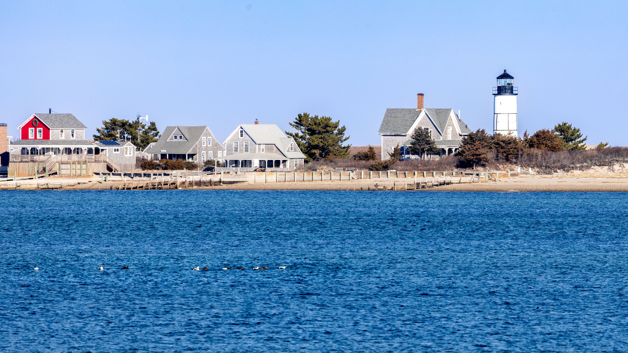 Picturesque Cape Cod lighthouse with traditional homes by the shoreline under a clear sky.