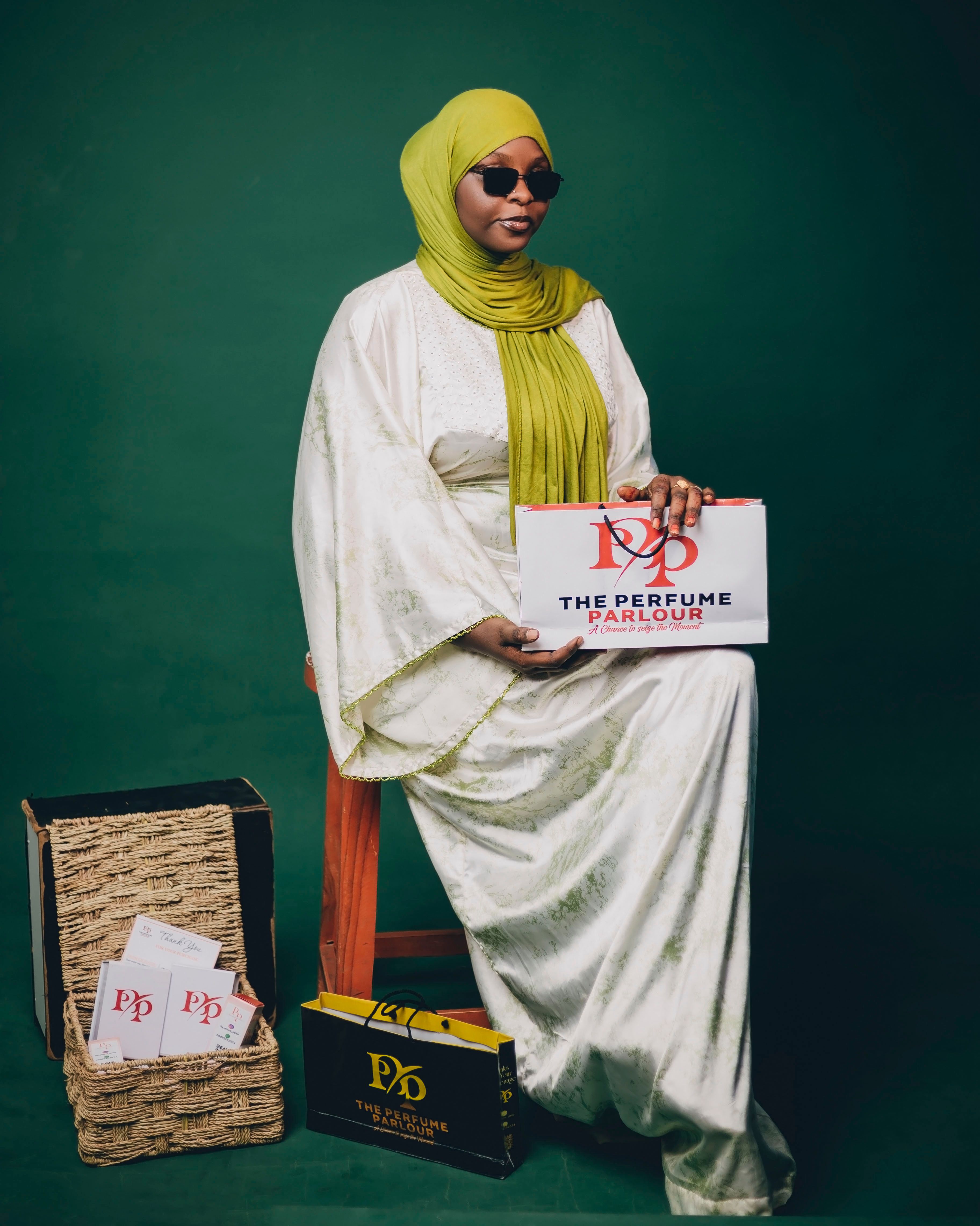 Elegant woman in a green hijab holding perfume packaging, seated indoors.
