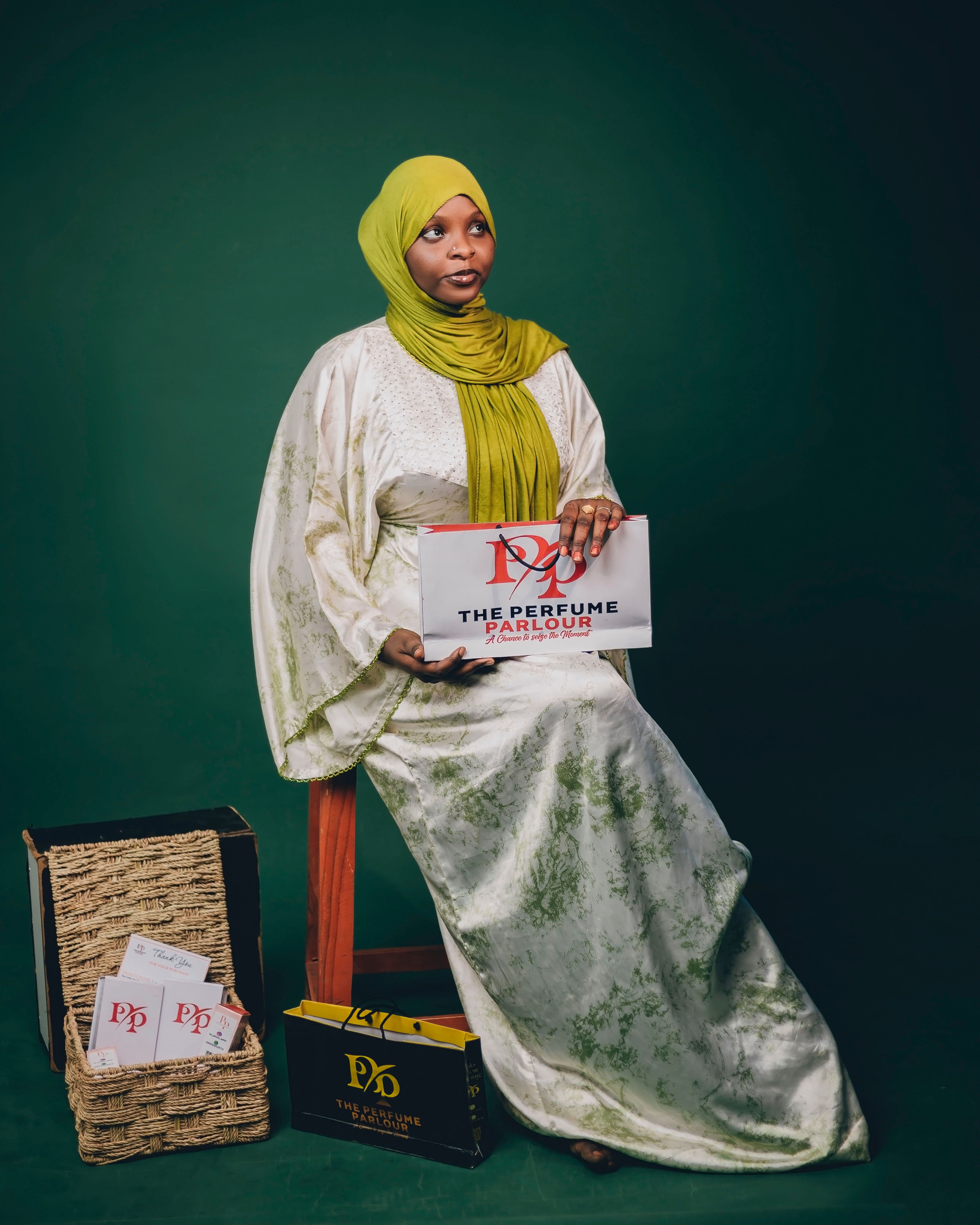 Woman in traditional attire holding a sign for The Perfume Parlour against green backdrop.