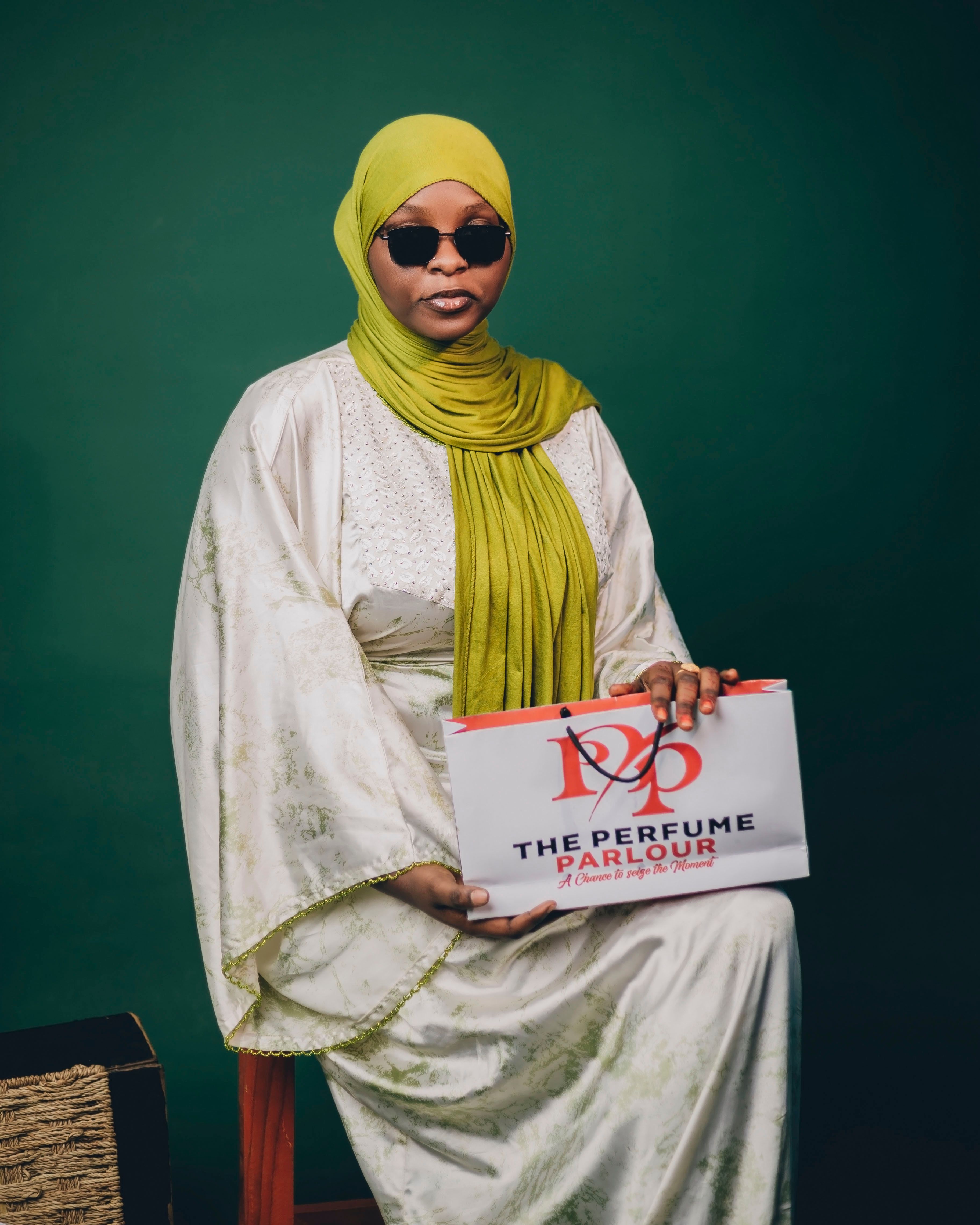 Elegant woman wearing green hijab and sunglasses, holding 'The Perfume Parlour' package against green backdrop.