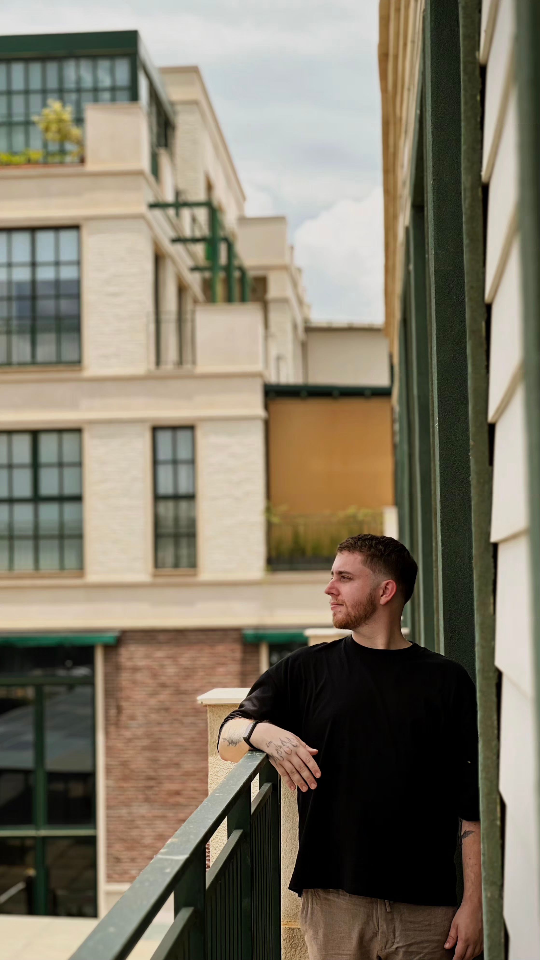 Young man in black shirt on balcony, looking thoughtfully at urban architecture.
