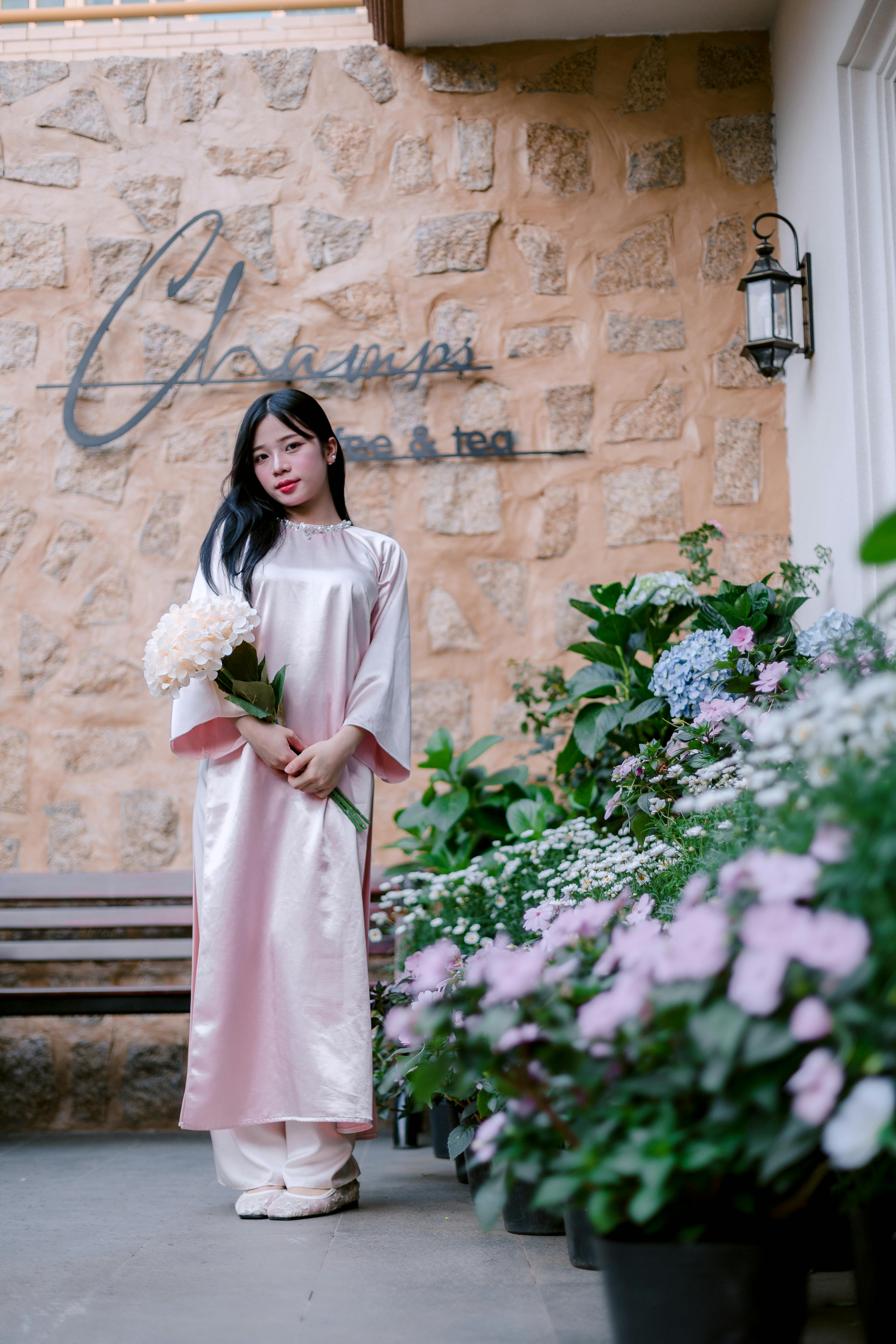 A young woman in a pink traditional dress stands elegantly by a flower garden.