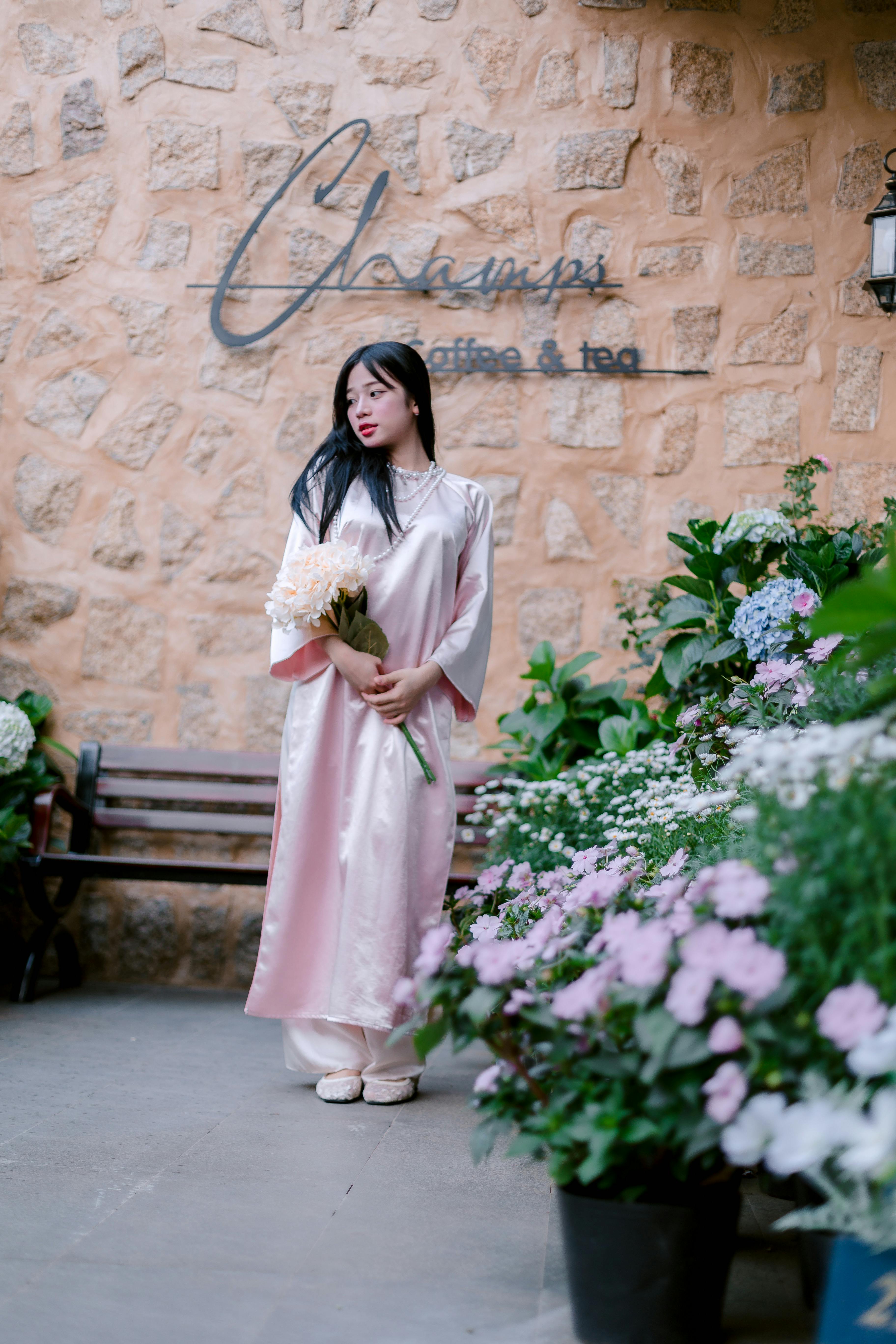Woman in a soft pink dress holding a bouquet at a café setting, surrounded by flowers.