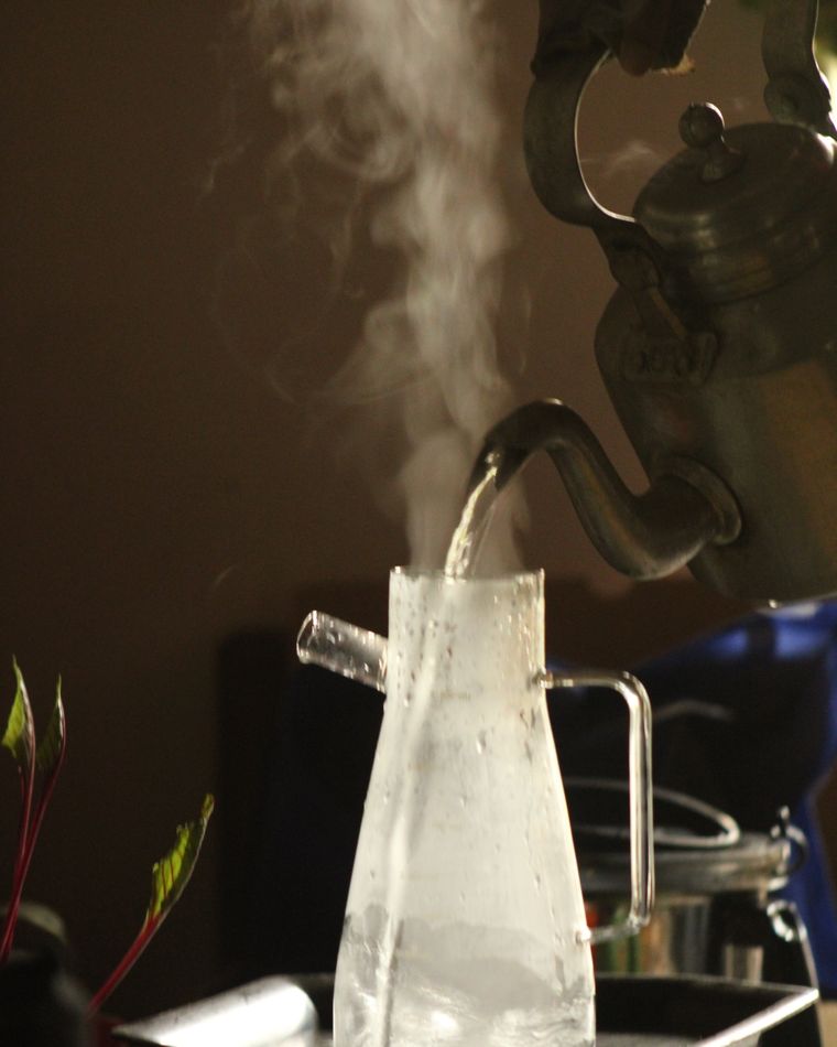 A kettle pouring hot water into a glass teapot in soft morning light.