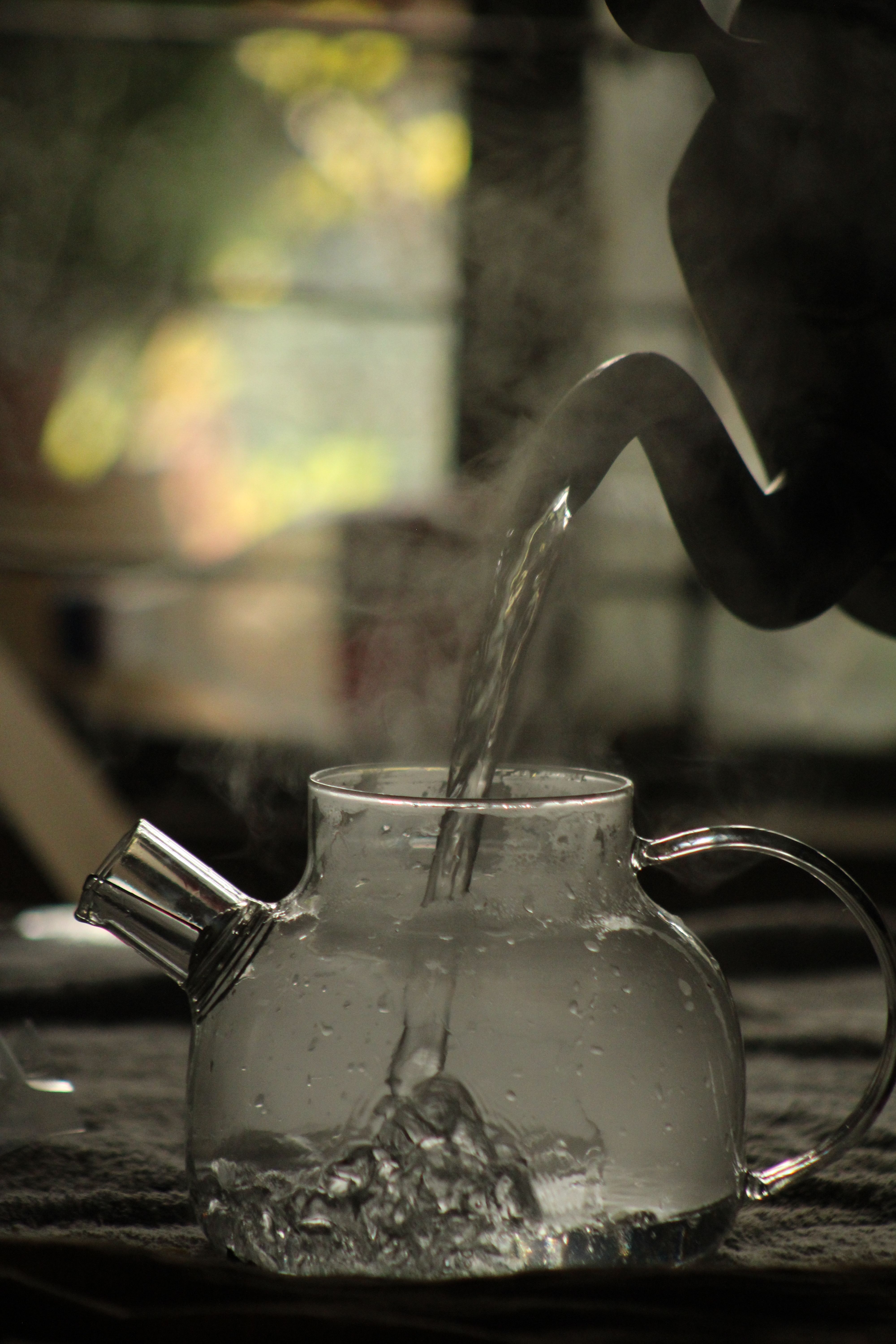 A side view of hot water being poured into a glass teapot with steam.