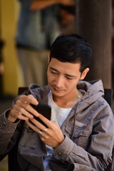 Young man in a jacket focused on his smartphone while seated outdoors in a relaxed setting.