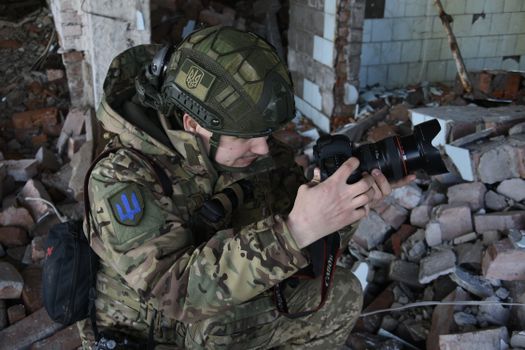 Military person in camouflage with camera amidst rubble, capturing images.