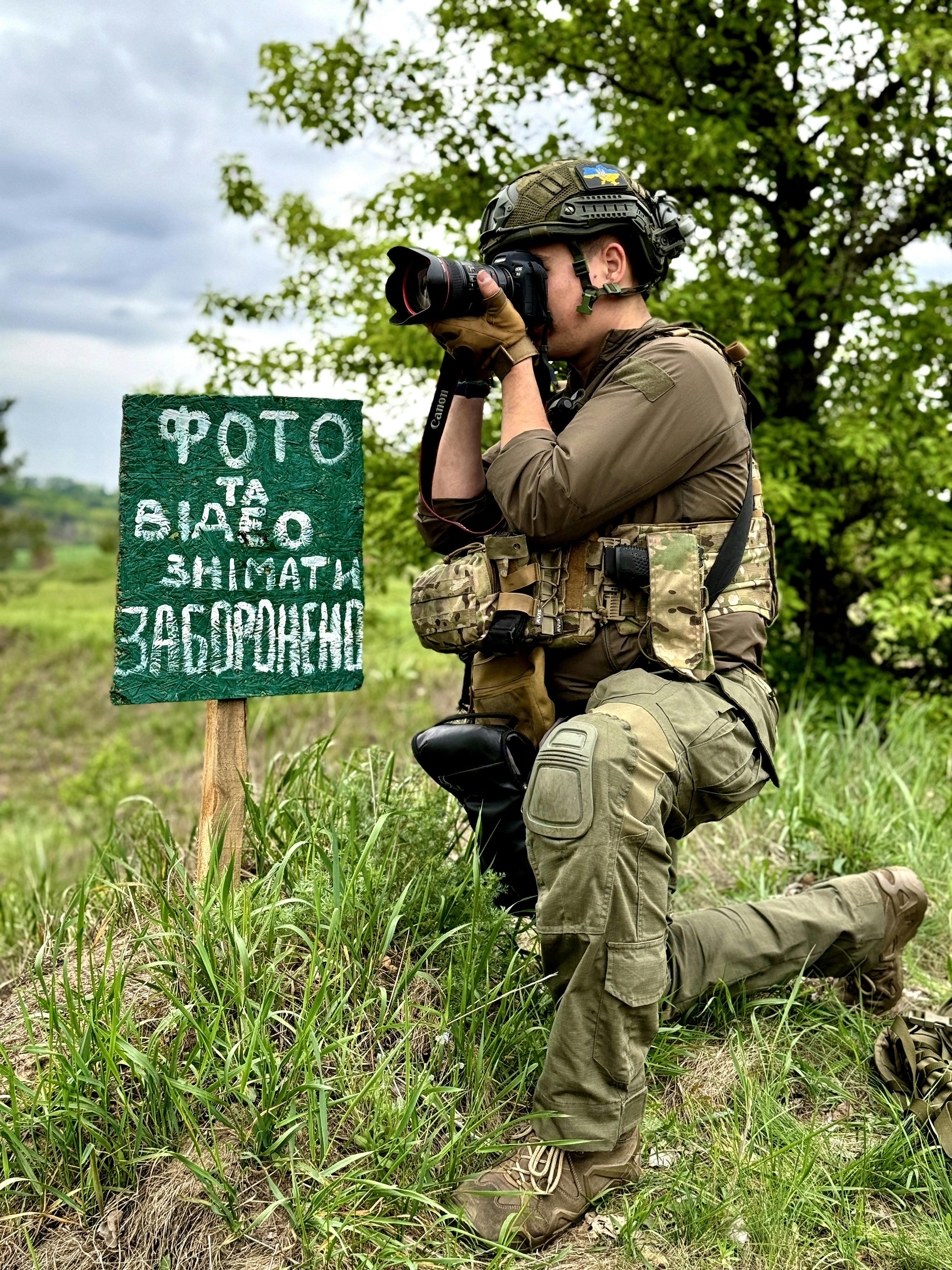 A military photographer defiantly takes a picture beside a sign that prohibits photography in a lush outdoor setting.