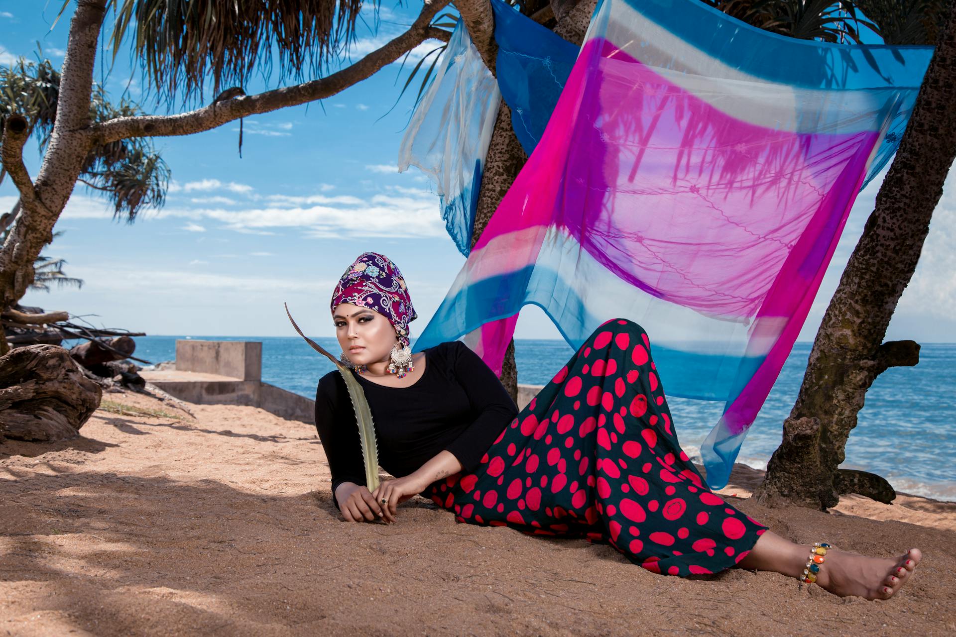 A stylish woman with a headscarf poses on a vibrant beach in Sri Lanka with colorful fabrics.