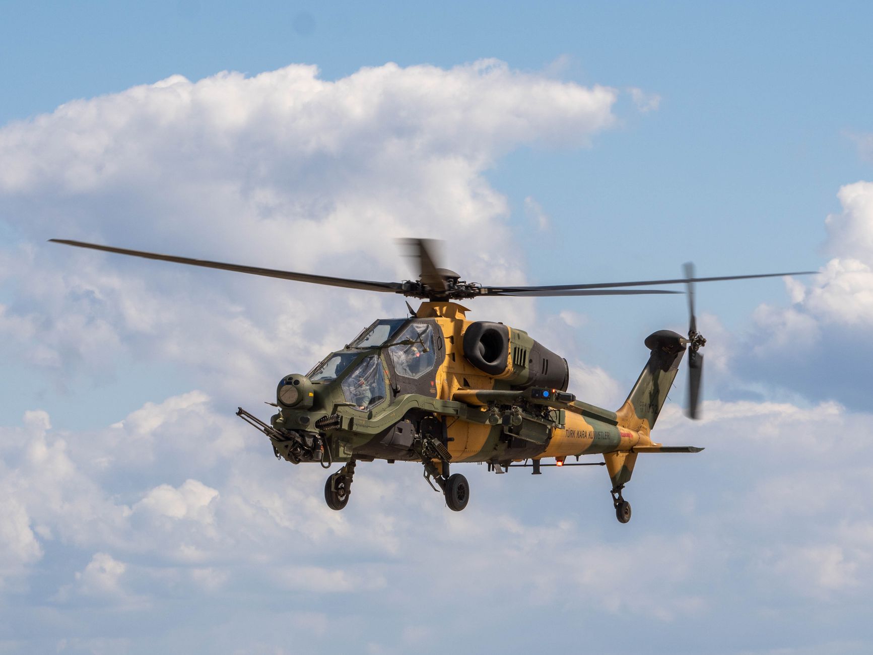 A camouflaged military helicopter flying against a backdrop of clouds on a clear day.