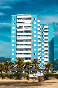 A modern blue high-rise building by the beach, set against a clear blue sky and palm trees.